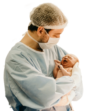 man wearing a white hair cap and white face mask and light blue scrubs holding an newborn child with his arms and his hands are gloved